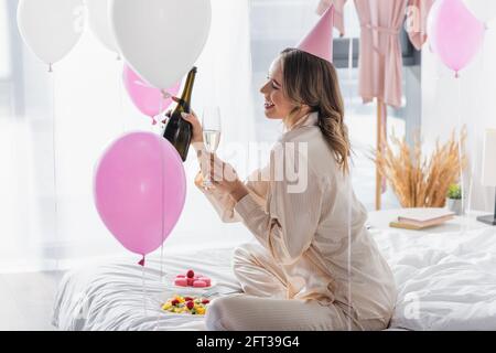 Side view of smiling woman holding glass and bottle of champagne near fruit salad and macaroons Stock Photo