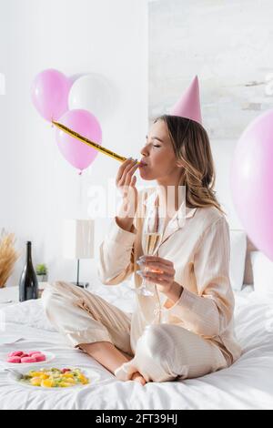 Woman blowing party horn and holding champagne near macaroons and fruit salad on bed Stock Photo