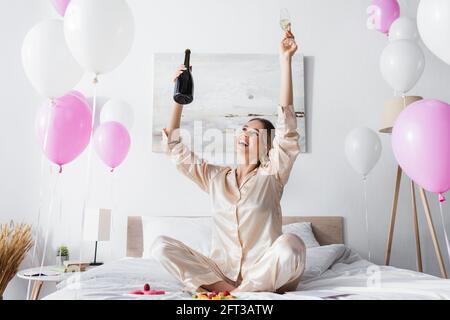 Smiling woman holding champagne near fruit salad, macaroons and festive balloons in bedroom Stock Photo