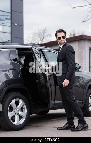 bearded bodyguard in suit and sunglasses with security earpiece opening car door