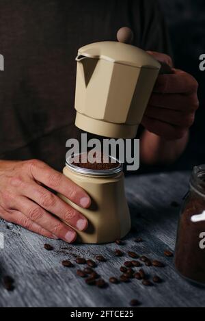 a young caucasian man prepares coffee in a beige moka pot, sitting at a gray rustic wooden table Stock Photo