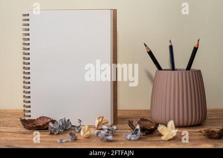 Open spiral notebook with white sheet, pencils in the glass next to it and decorations on the table Stock Photo