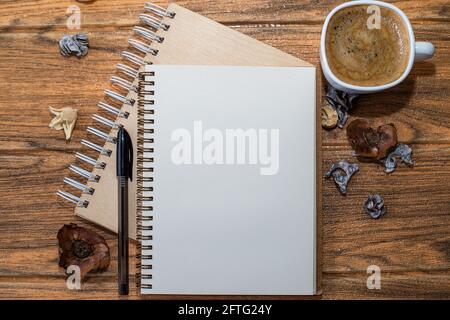 Spiral notebooks, one open with white sheet and black pen next to it. On the wooden table there is another cup of coffee and decorations. Stock Photo