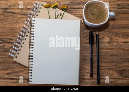 Spiral notebooks with 3 flowers. One open with white sheet and black pen next to it. There is another cup of coffee on the wooden table. Stock Photo