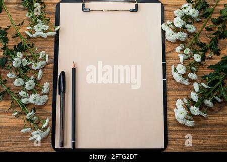Black clipboard with ivory sheet, on it are a pen and a pencil. There are white flowers on both sides of the clipboard. Stock Photo