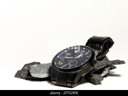 Dark wristwatch lays on top of a pile of dark rocks on a white background Stock Photo