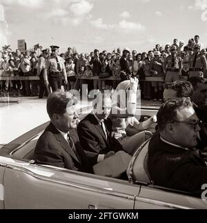 President John F. Kennedy and First Lady Jacqueline Kennedy ride in the back seat of a convertible as their motorcade travels en route to the Orange Bowl Stadium in Miami, Florida, for a presentation ceremony of the 2506th Cuban Invasion Brigade flag. President Kennedy holds in his lap a large envelope inscribed with a handwritten notation that reads, 'Speech - Miami'; Mayor of Miami, Robert King High, sits between the President and Mrs. Kennedy (wearing white gloves and a head scarf wrapped under her chin). In front seat of car (left to right): White House Secret Service agent, Floyd Boring. Stock Photo