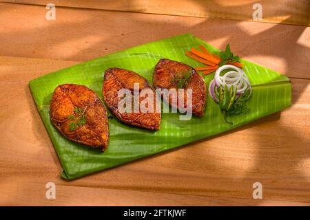 Seer fish fry arranged beautifully and garnished with onion, lemon and tomato slices on banana leaf covered base which is placed on wooden texture wit Stock Photo