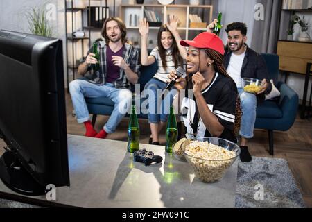 Happy three friends sitting on couch and dancing with hands while african woman singing in wireless microphone. Karaoke time during home party. International friendship. Stock Photo