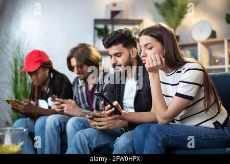 Male and female friends sitting together on sofa and ignoring each other while using personal smartphones. Young diverse people obsessed with modern gadgets. Stock Photo