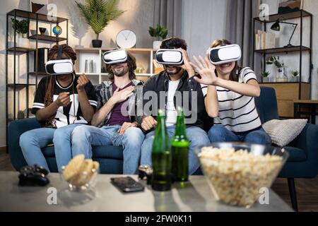Group of four multi ethnic friends using wireless headset for experiencing virtual reality. Joyful men and women sitting on comfy couch and playing playing games. Technology concept. Stock Photo