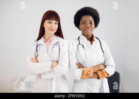 Two smiling happy African and Caucasian female doctors colleagues, wearing white coats, standing side by side with folded arms, posing at camera at modern light medical office Stock Photo