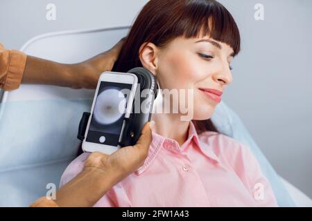 Mole checkup. Professional dermatoscopy. Close-up of dermatologist hands checking face skin of charming young 30-aged European lady with digital photo dermatoscope Stock Photo