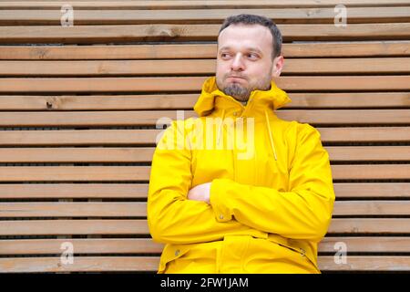 young European man with sly look, beard sitting outside on wooden bench in yellow jacket with crossed hands, blowing cheeks, looking aside, cunning offended, thinking over something, keeping a secret Stock Photo