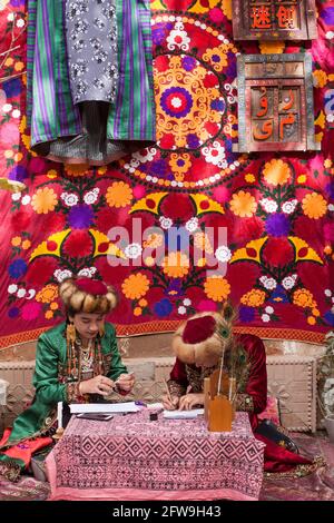 Two young women in colorful ethnic dresses sitting writing in arabic. Kashgar, Xinkiang, Popular Republic of China, 2019 Stock Photo