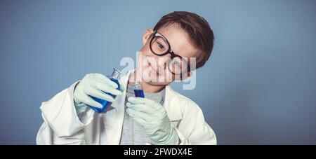 cool school boy with thick black glasses is dressed as scientist with white coat and experimenting with blue liquids in front of blue background Stock Photo