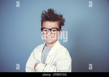 cool school boy with thick black glasses is dressed as mad scientist with white coat in front of blue background Stock Photo