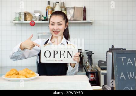 Young asian shopkeeper with a smile holds an OPEN sign in front of a coffee shop counter. Morning atmosphere in a coffee shop. Stock Photo