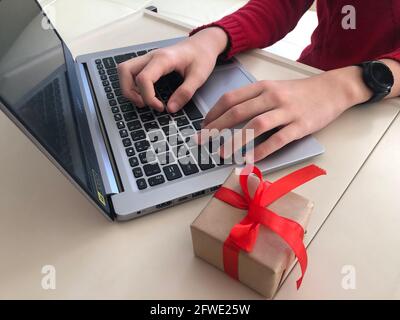 Gift box decorated with a red ribbon bow in hands against the background of the office workplace, gift delivery, coffee break during working hours, co Stock Photo