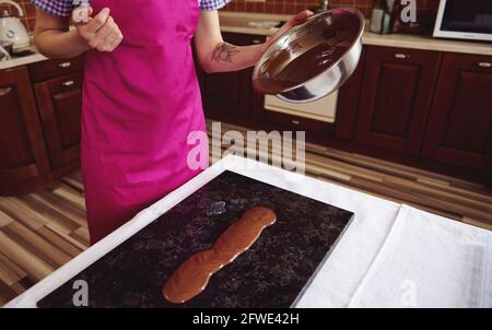 Female confectioner chocolatier wearing pink apron pouring melted chocolate on marble surface on the background of wooden kitchen