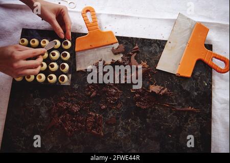 Flat lay composition with confectioner hands putting chocolate filling into spherical molds from white chocolate. Tempered chocolate mass and scrabers Stock Photo