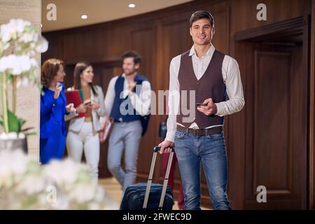 group of young business people, walking through the hallway. Selective focus on a male with cell phone, carrying wheeled suitcase. Stock Photo