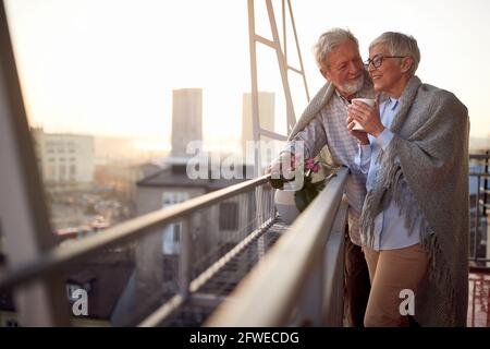 An elderly couple is in romantic moments while they enjoying the view on the city from the terrace of their apartment at a beautiful sunset. Spouses, Stock Photo