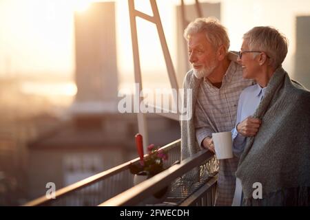 An elderly couple enjoying a view on the city from the terrace of their apartment at a beautiful sunset. Spouses, pensioners, together, home Stock Photo