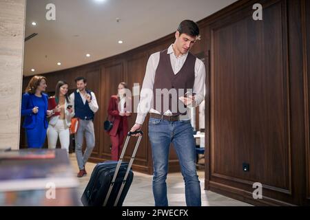 young businessman pulling suitcase on wheels, looking at his cellphone with colleagues walking behind him Stock Photo