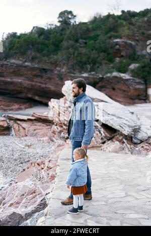 Dad in a denim suit holds the hand of a little girl in a dress and a denim jacket while standing on a rocky coast against a background of mountains Stock Photo