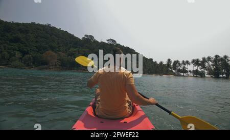 Man with sunglasses and hat rows pink plastic canoe along sea against green hilly islands with wild jungles. Traveling to tropical countries. Stock Photo