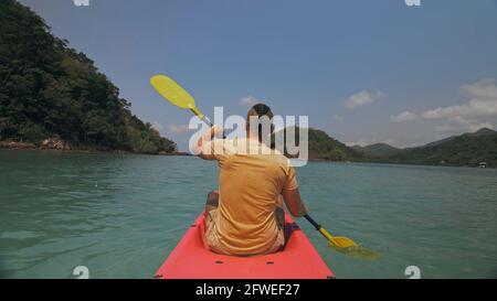 Man with sunglasses and hat rows pink plastic canoe along sea against green hilly islands with wild jungles. Traveling to tropical countries. Stock Photo