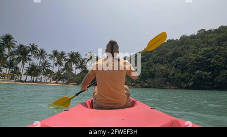 Man with sunglasses and hat rows pink plastic canoe along sea against green hilly islands with wild jungles. Traveling to tropical countries. Stock Photo