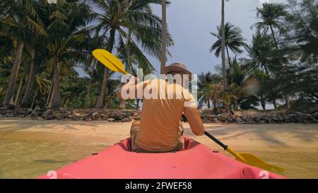 Man with sunglasses and hat rows pink plastic canoe along sea against green hilly islands with wild jungles. Traveling to tropical countries. Stock Photo