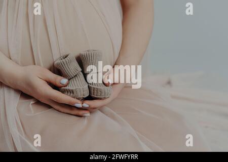 Unrecognizable pregnant woman holds small boots over tummy wears festive dress. Faceless future mother Stock Photo
