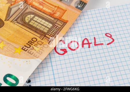 Word GOALS is written in red on a checkered paper notepad. Fifty euro banknote opens an inscription close up. Wealth goal definition and achievement c Stock Photo