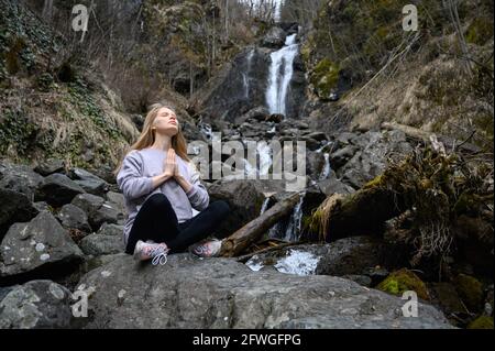 Girl sits on a large stone and meditates near a waterfall next to a mountain river. Stock Photo