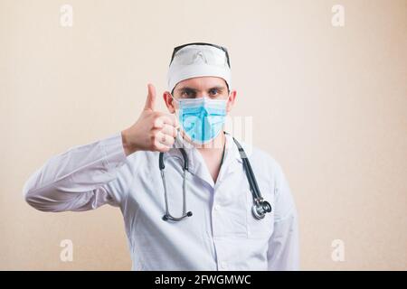 Young doctor portrait in white suit and protective breathing mask glasses and stethoscope with thumb up isolated Stock Photo
