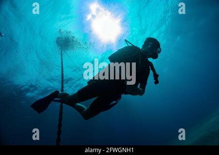 Silhouette of young diver against Sun light at the sea surface and hanging down steel anchor chain. Air bubbles from breathing. Scuba diving in tropic Stock Photo