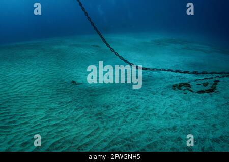 Underwater photo of heavy steel anchor chain that hangs down from water surface and lays on white sand bottom, murky blue water deeper in tropical sea Stock Photo