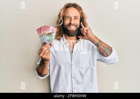 Handsome man with beard and long hair talking on the phone holding 100 new zealand dollars smiling and laughing hard out loud because funny crazy joke Stock Photo