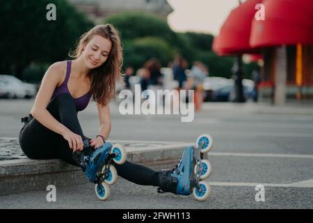 Outdoor shot of young sporty woman in sportswears puts on roller blades poses on road going to ride rollers during sunny day ties shoelaces has good Stock Photo