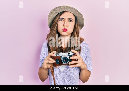 Young hispanic girl wearing summer hat holding vintage camera puffing cheeks with funny face. mouth inflated with air, catching air. Stock Photo