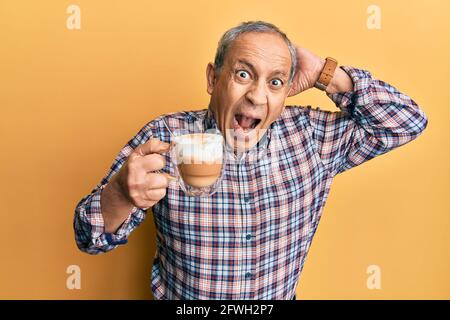 Handsome senior man with grey hair drinking a cup coffee crazy and scared with hands on head, afraid and surprised of shock with open mouth Stock Photo