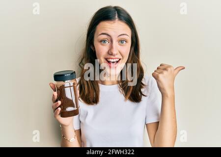 Young brunette woman holding soluble coffee pointing thumb up to the side smiling happy with open mouth Stock Photo