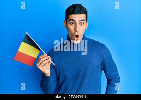 Handsome hispanic man holding belgium flag scared and amazed with open mouth for surprise, disbelief face Stock Photo