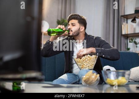 Stylish indian guy drinking cold beer while sitting comfortably on couch and enjoying football championship at home. Concept of free time, sport and entertainment. Stock Photo