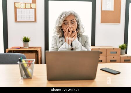 Middle age businesswoman sitting on desk working using laptop at office shocked covering mouth with hands for mistake. secret concept. Stock Photo