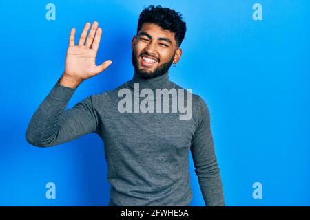 Arab man with beard wearing turtleneck sweater waiving saying hello happy and smiling, friendly welcome gesture Stock Photo
