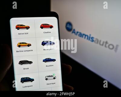 Person holding smartphone with webpage of French online car dealer Aramis SAS (Aramisauto) on screen with logo. Focus on center of phone display. Stock Photo